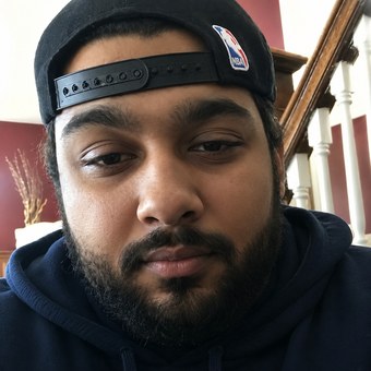 Photograph of Joseph Tennis wearing a dark backwards baseball cap and a dark hooded top, indoors, cropped to show him alone.